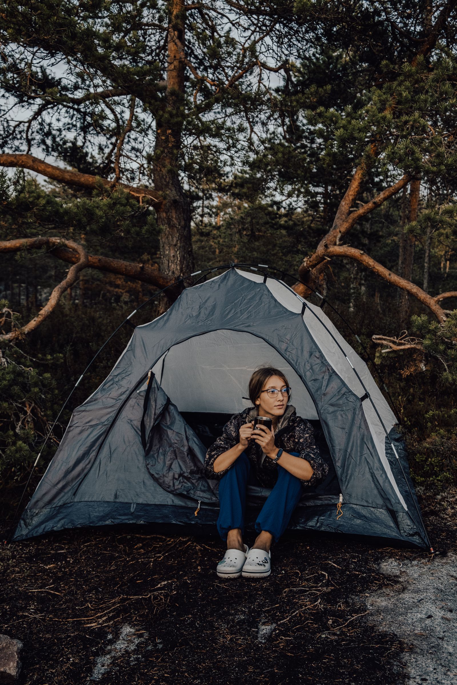 A woman sits in a tent doorway with a mug, pine forest behind her.