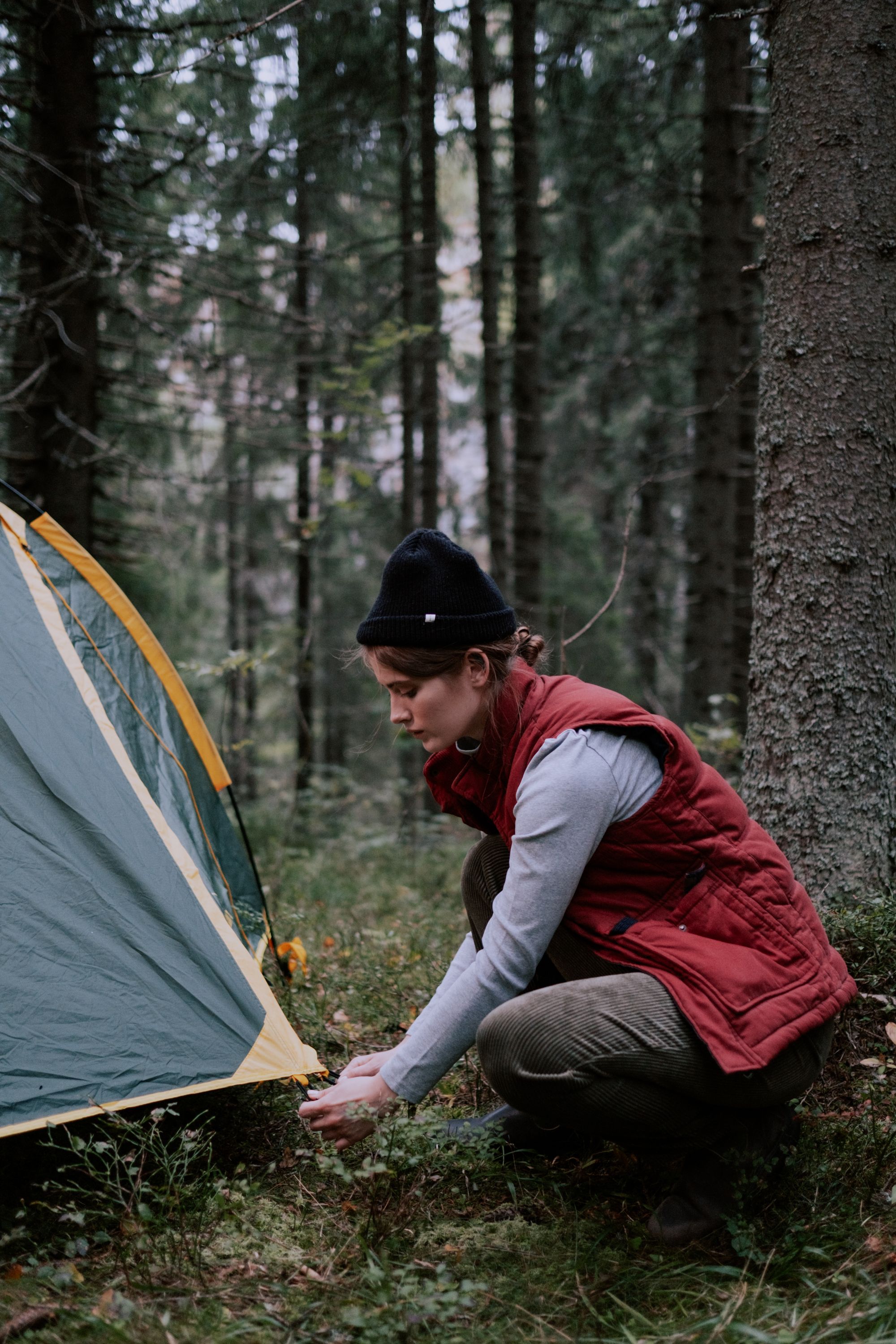 A woman in a rust vest stakes a green tent on a mossy forest floor.